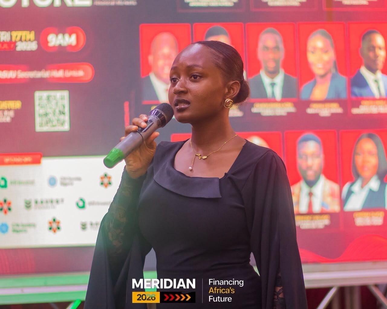 Maryjane Ejebughaobi hosting on stage with a microphone in front of a speaker wall